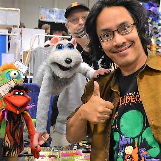 A man in glasses gives a thumbs up beside a gray mouse puppet and other puppets at a convention booth