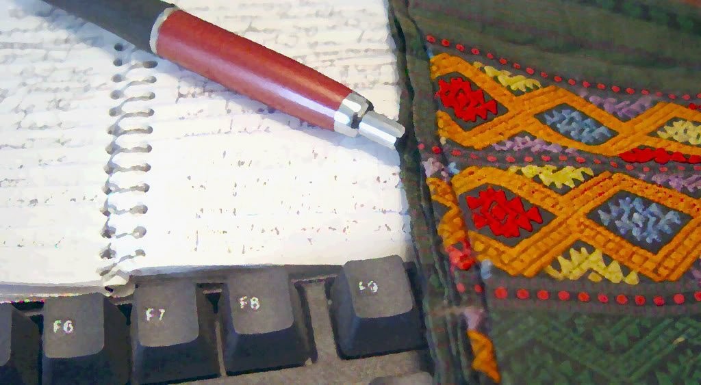 Handwritten notebook and pen on a keyboard beside a woven Lao textile in red, gold, and blue