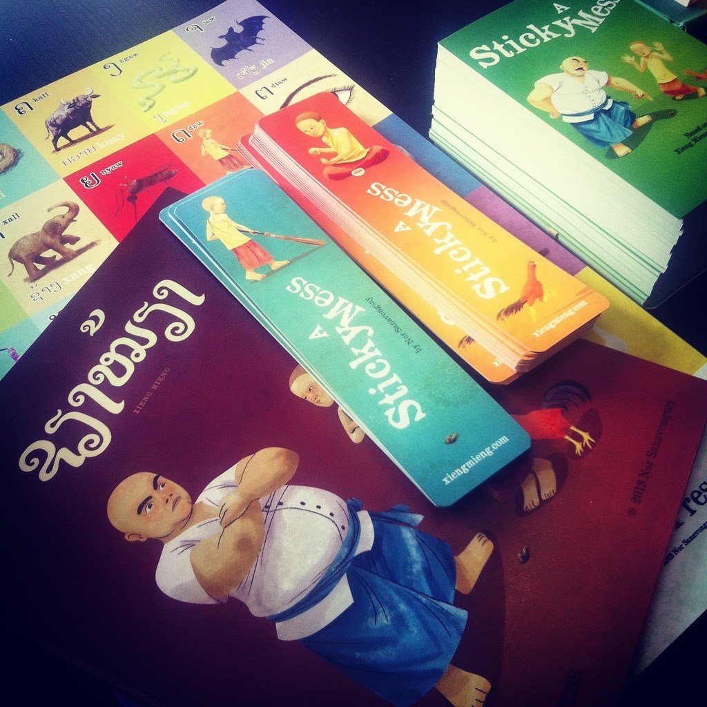 A Sticky Mess books, bookmarks, and a Lao alphabet poster spread on a table, with the Xieng Mieng edition in Lao script
