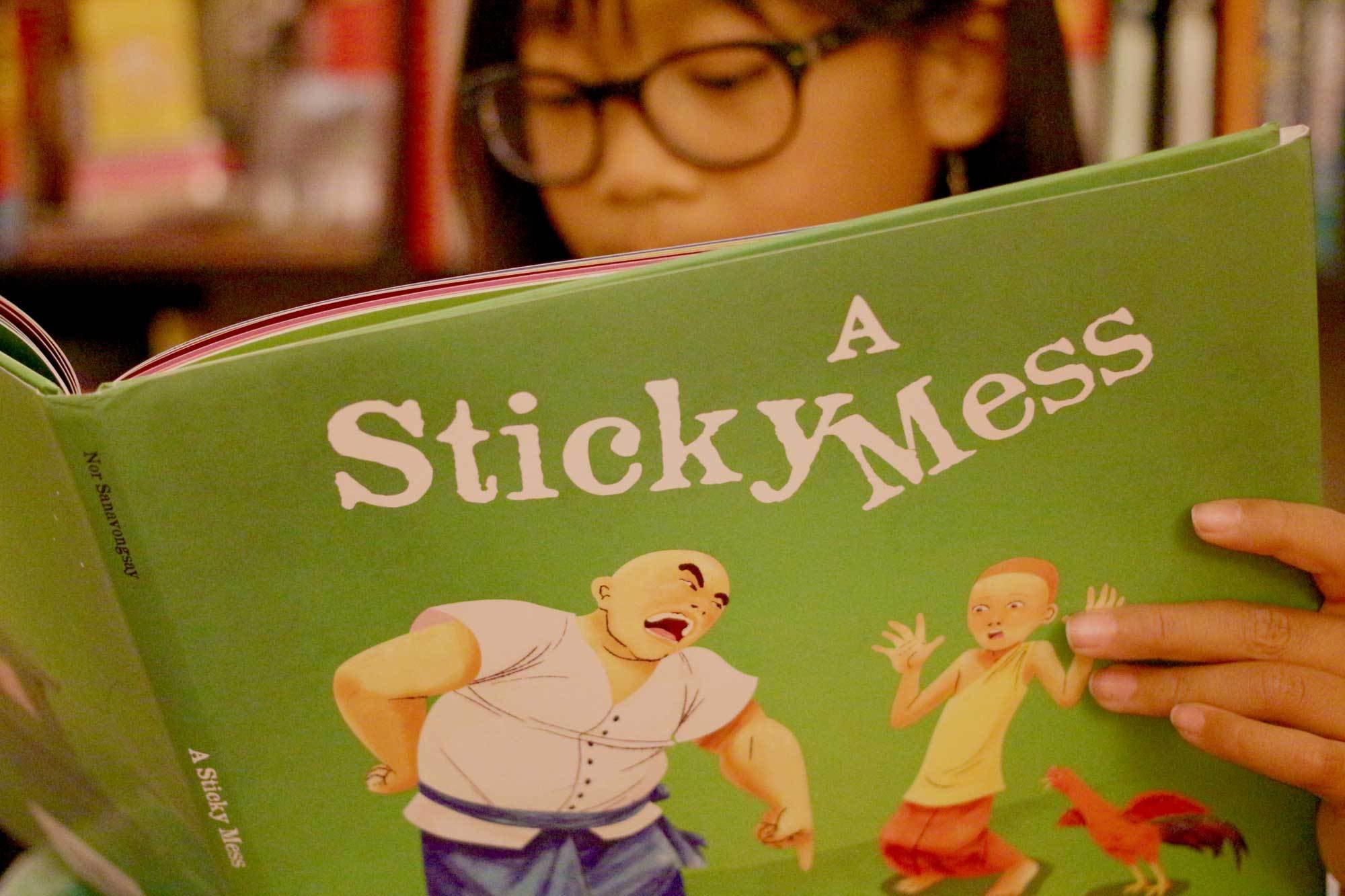 Child in glasses reading A Sticky Mess, its green cover showing an angry bald man, a young novice and a rooster
