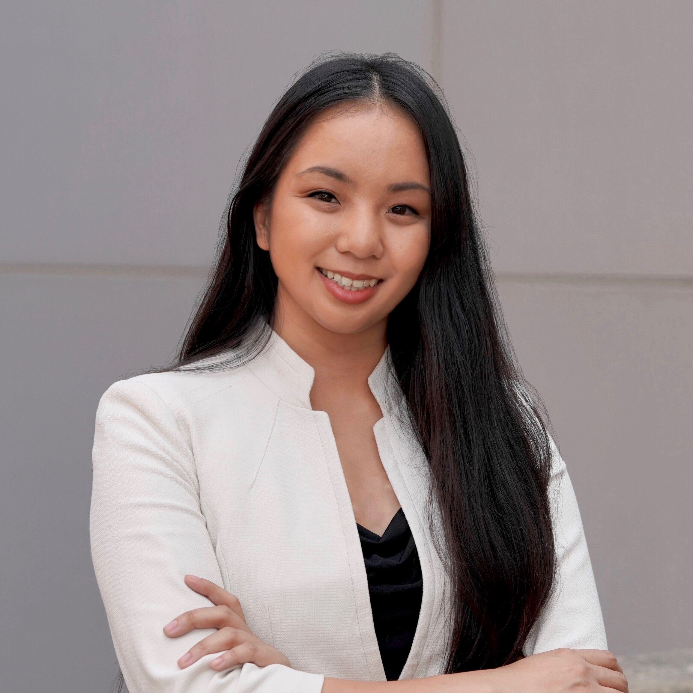 Portrait of Mai Nguyen Do smiling, with long black hair, in a white blazer over a black top against a gray wall.