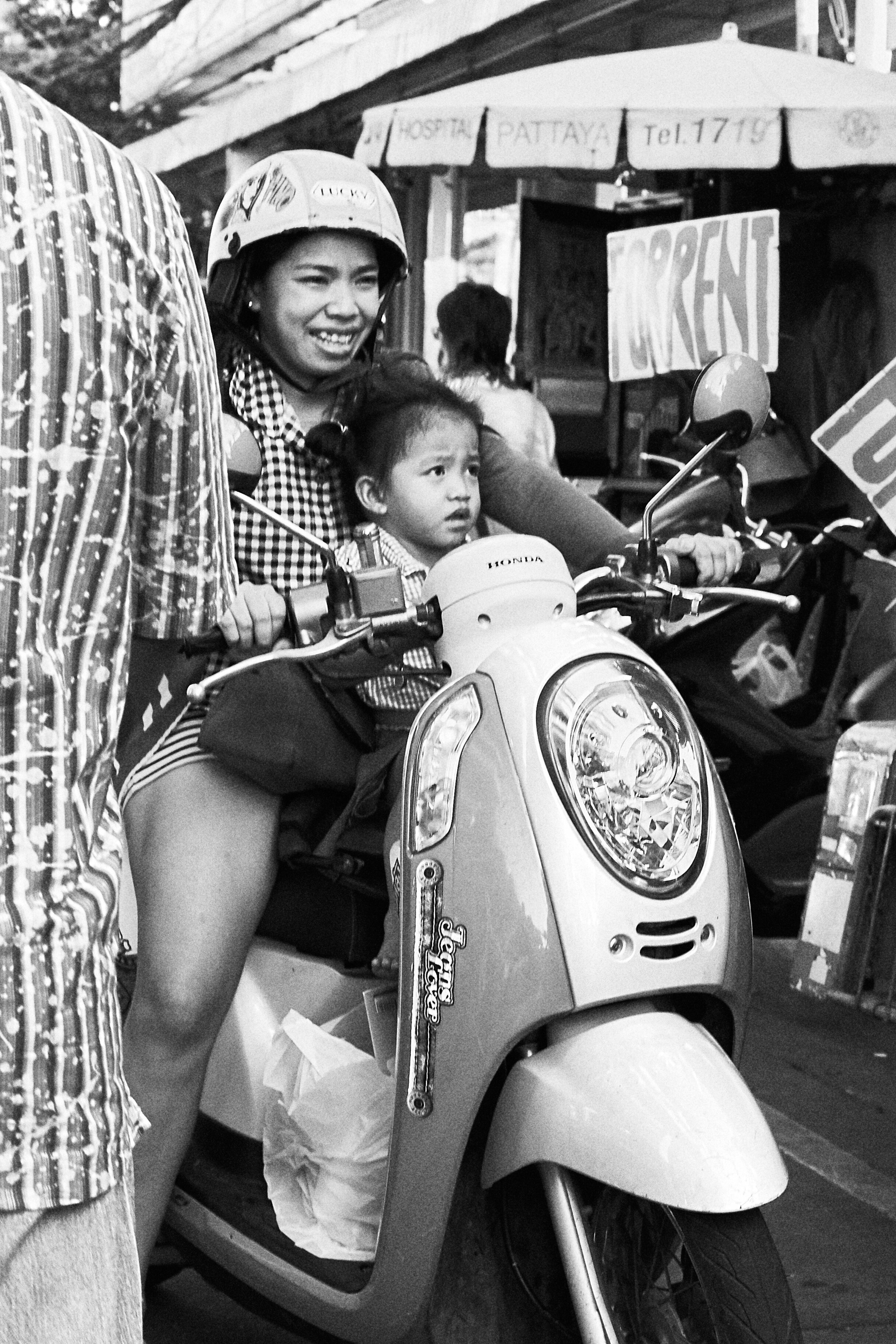 A woman and young girl sitting on a Honda scooter, with the woman smiling and wearing a helmet. The girl has a serious expression. They are in a busy outdoor setting with signs and market stalls in the background.