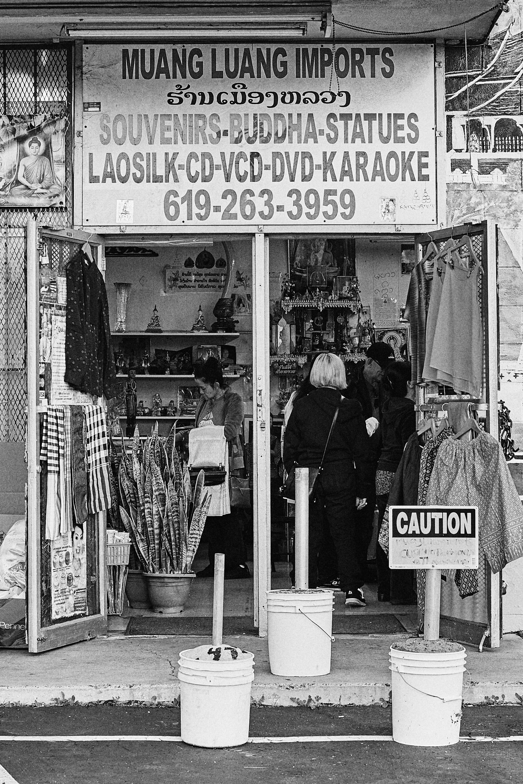 A storefront with a large sign reading 'Muang Luang Imports,' featuring items like souvenirs, Buddha statues, CDs, VCDs, DVDs, and karaoke, with people shopping inside and buckets on the sidewalk.