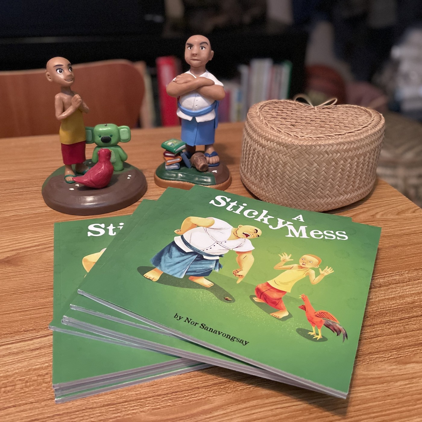 Stack of green children's books titled 'A Sticky Mess' by Nor Sanavongsay on a wooden table, surrounded by small figurines including a boy with crossed arms, a boy praying, a green dog, and a bird, with a woven basket and a bookshelf in the background.