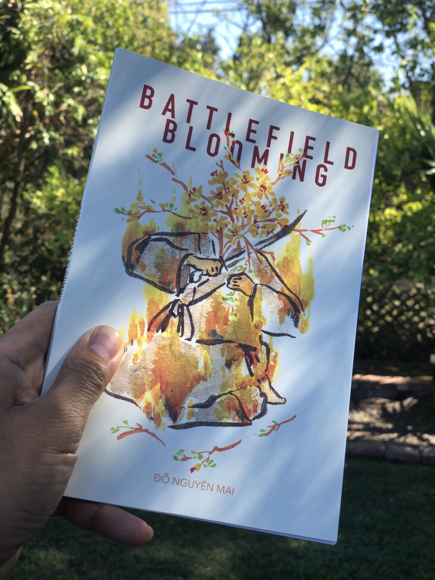 Hand holding a copy of Battlefield Blooming by Đỗ Nguyên Mai outdoors in a sunny garden