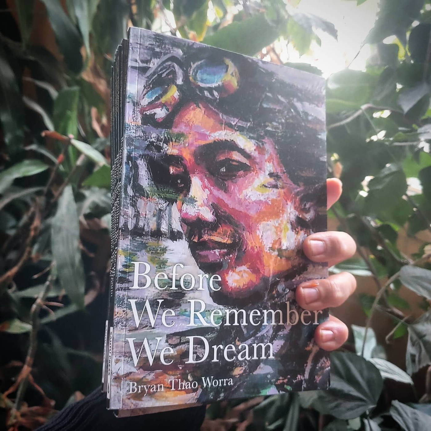 Hand holding a copy of Before We Remember We Dream by Bryan Thao Worra among green leaves