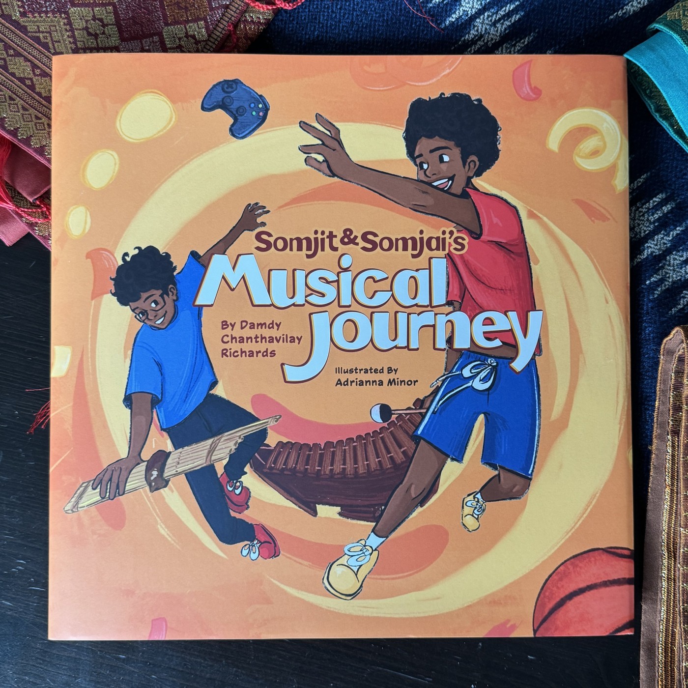 Book cover titled "Somjit & Somjai's Musical Journey" featuring two children, one playing a traditional instrument and the other jumping with a tambourine, set against a vibrant background with musical instruments and abstract swirls.